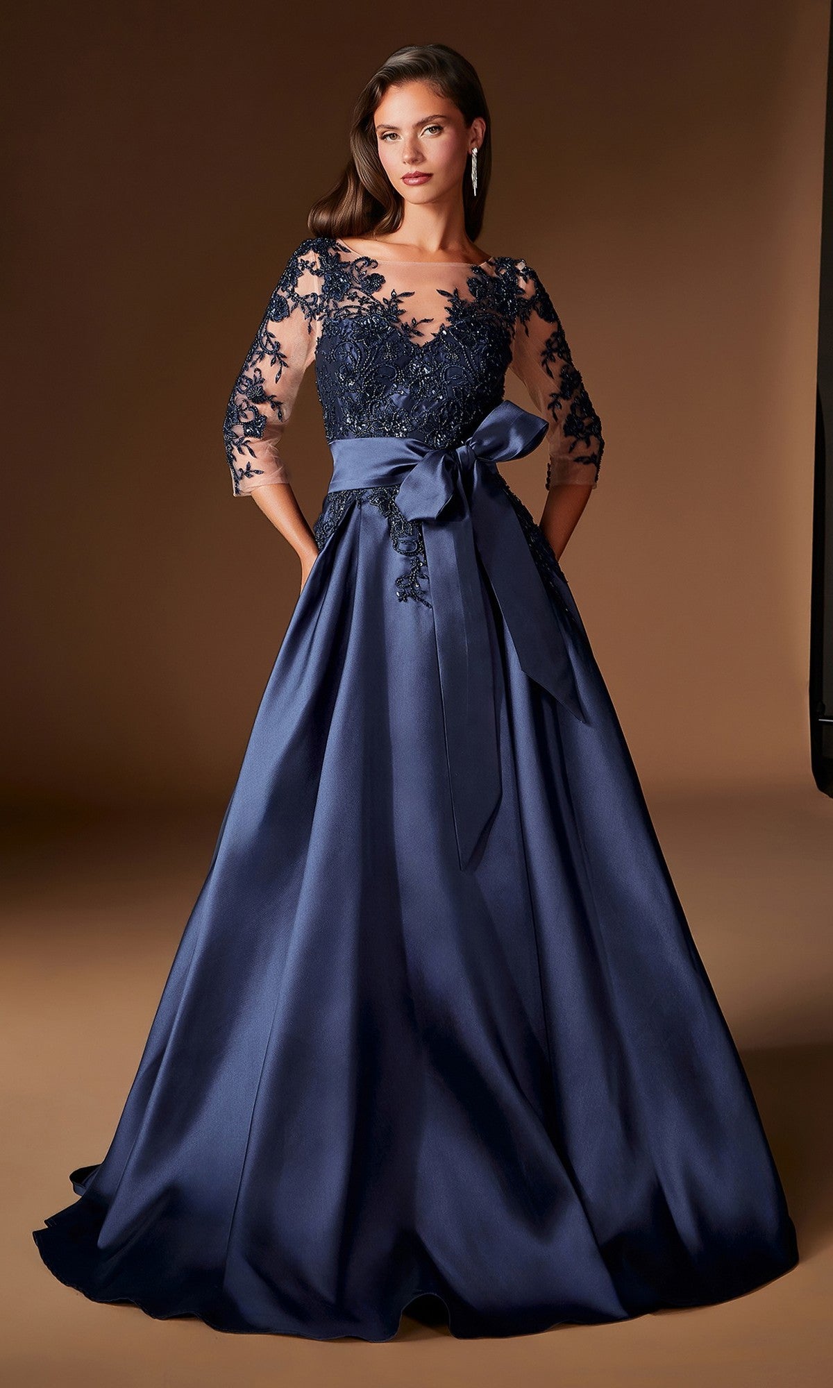 A woman wearing the Ladivine CDS569 Long Formal Dress—a floor-length, dark blue evening gown featuring a lace bodice, sheer sleeves, and a satin bow at the waist—stands against a neutral brown background.